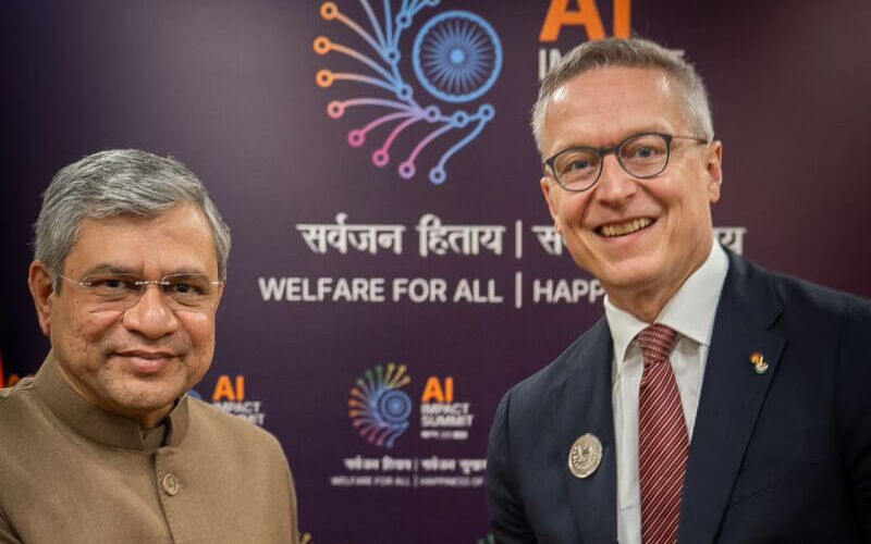 Zwei Männer in formeller Kleidung schütteln sich die Hand bei einem offiziellen Treffen vor einer Wand mit der Aufschrift ‚AI Impact Summit‘