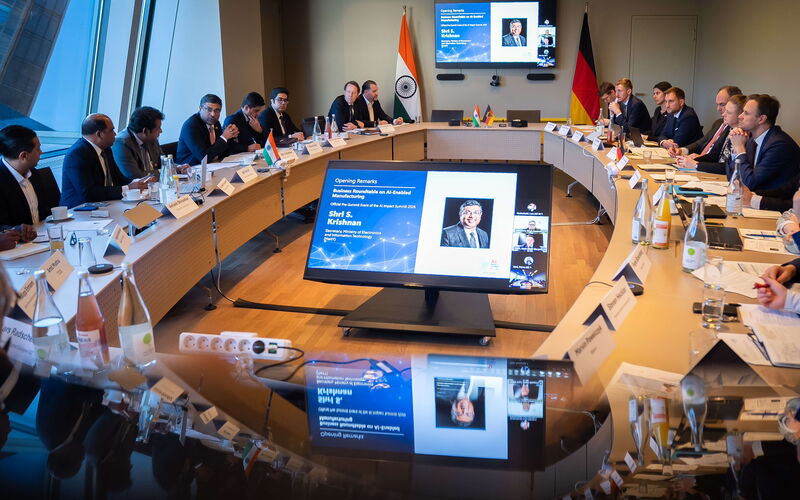 Teilnehmende aus Regierung, Wirtschaft und Forschung sitzen an einem großen Konferenztisch beim Indo-German Digital Dialogue und verfolgen eine Präsentation zu KI-gestützter Industrie, die auf mehreren Bildschirmen angezeigt wird.