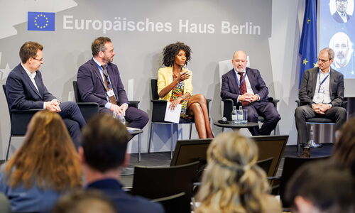 Vier Diskussionsteilnehmer und der Moderator sitzen in einem Halbkreis auf der Bühne, und der Moderator in der Mitte hält die Einführungsrede zur Veranstaltung.