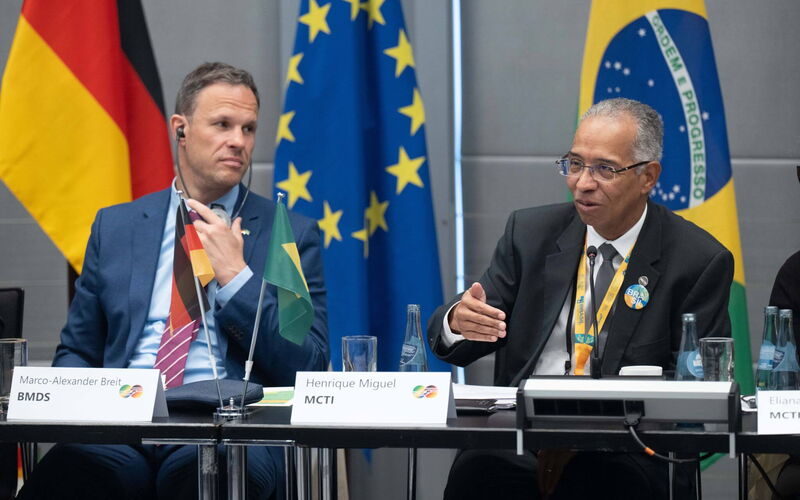 Das Foto zeigt Henrique Miguel vom MCTI und Marco-Alexander Breit vom BMDS vor einer deutschen, einer brasilianischen und einer europäischen Flagge.